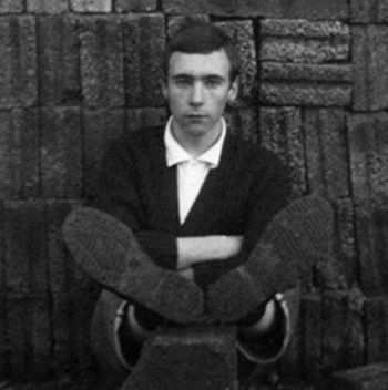 Black and white photo  of young  Martin Lovis sitting on the ground in front of a brick wall with his feet wearing boots raised up on a brick showing the soles of his boots.