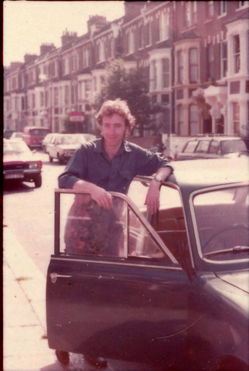 Colour photo of Martin Lovis standing next to a green car in the street outside his home in London 1982