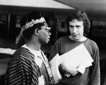 Black and white photo of Martin Lovis in discussion with a male dressed in traditional african wear visiting one of Lovis's exhibitions.