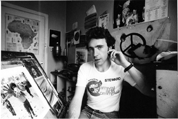 Black and white photograph of Martin sitting in front of his easel and surrounded by studio equipment with various posters and artwoks on the walls.