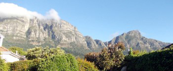 Bright sunlit distant view of Table Mountain, east side, as the cloud table cloth is being laid over the buttresses with Devil's Peak clearly shown tall and on its own with misty cloud slightly covering its peak.