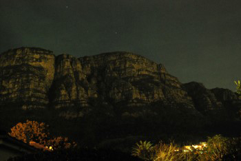 Moonlight reflected on buttresses of Table Mountain with red and white stars above and tiny house in the distance at the foot of the mountain with their house lights on.