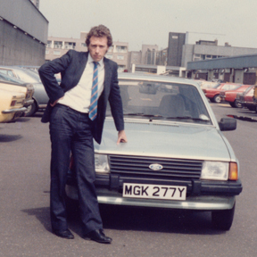 Colour photograph of Martin Lovis wearing a pin-stripe  business suit and tie leaning on the bonnet of his company car. London, 1985